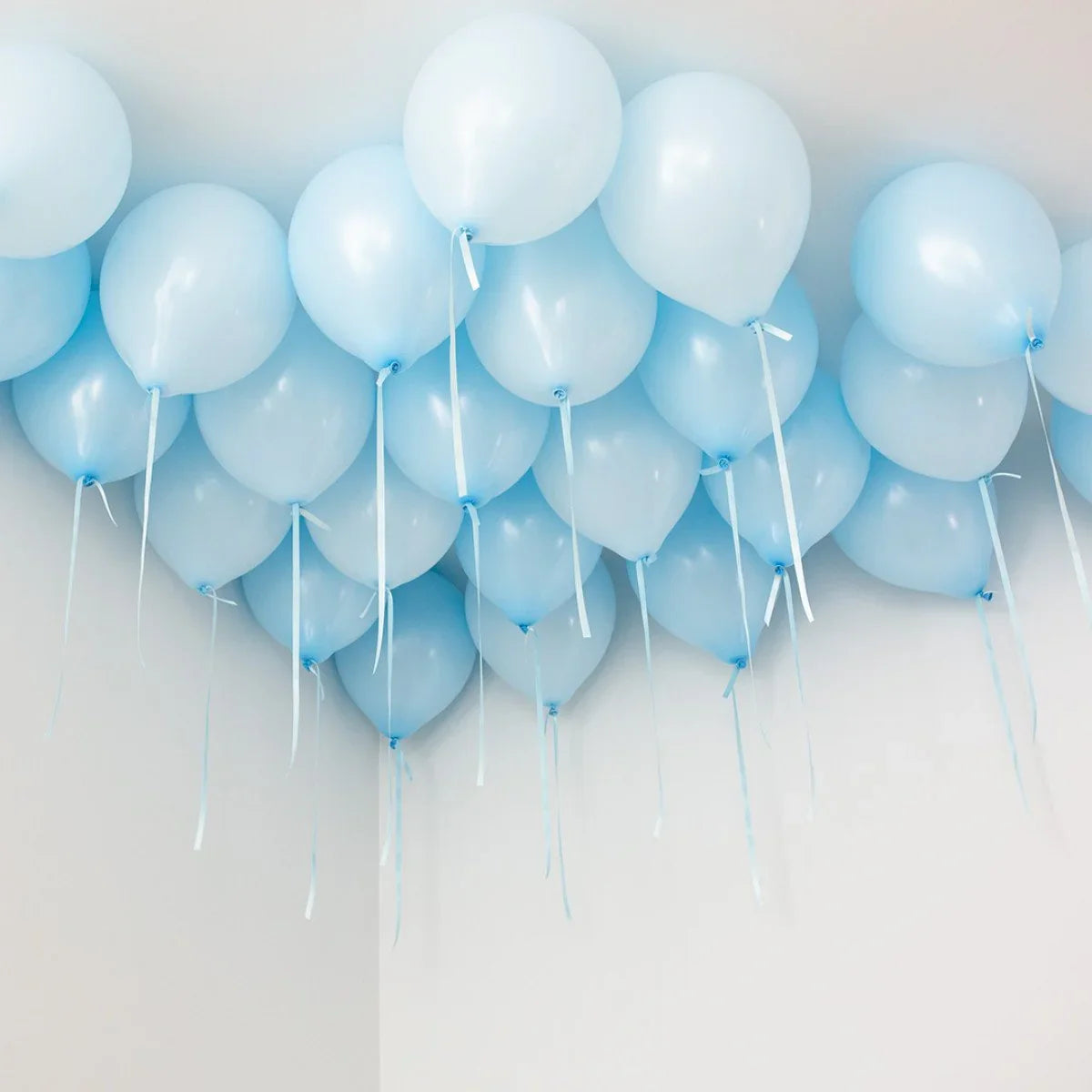 Cluster of baby blue balloons with ribbons floating on a white ceiling in a minimalist room