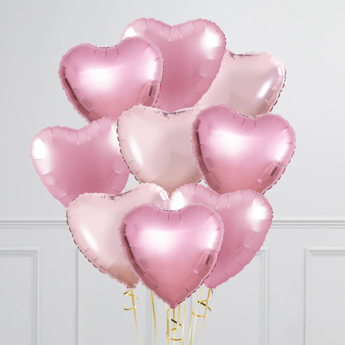 Bunch of shiny baby pink and light pink heart-shaped foil balloons with golden ribbons against a white wall