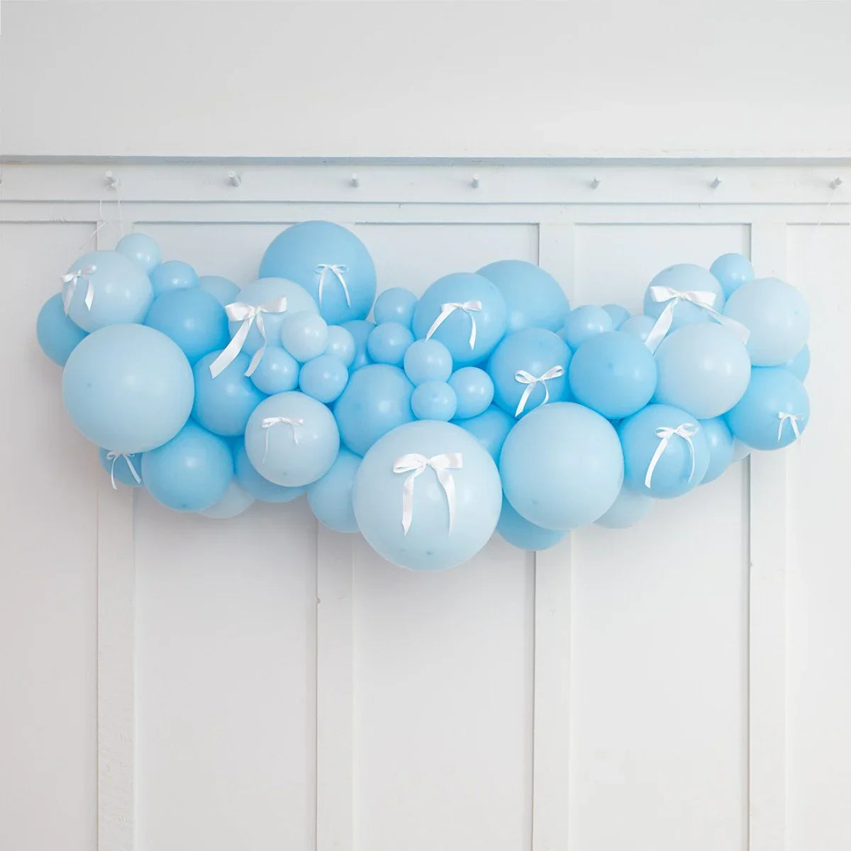 Blue balloon garland with white ribbon bows on white paneled wall, baby shower decoration