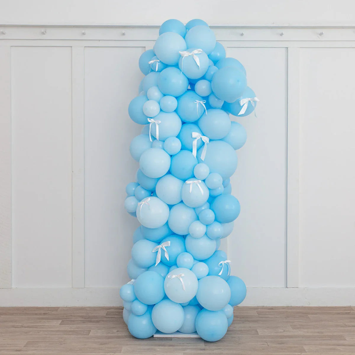 Tall blue balloon pillar decorated with white ribbons against white paneled wall and wooden floor