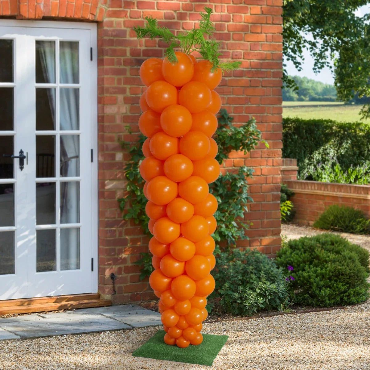 Large orange balloon sculpture shaped like a carrot with green balloon leaves, outdoor garden setting