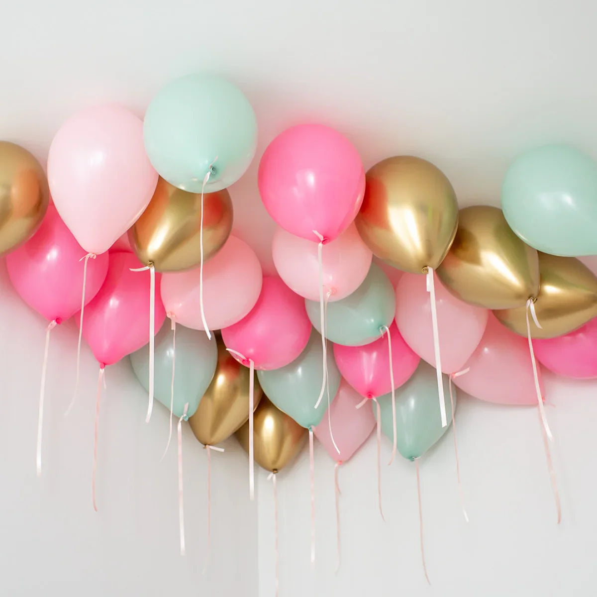 Cluster of helium balloons in pastel pink, mint green, and metallic gold floating on a white ceiling