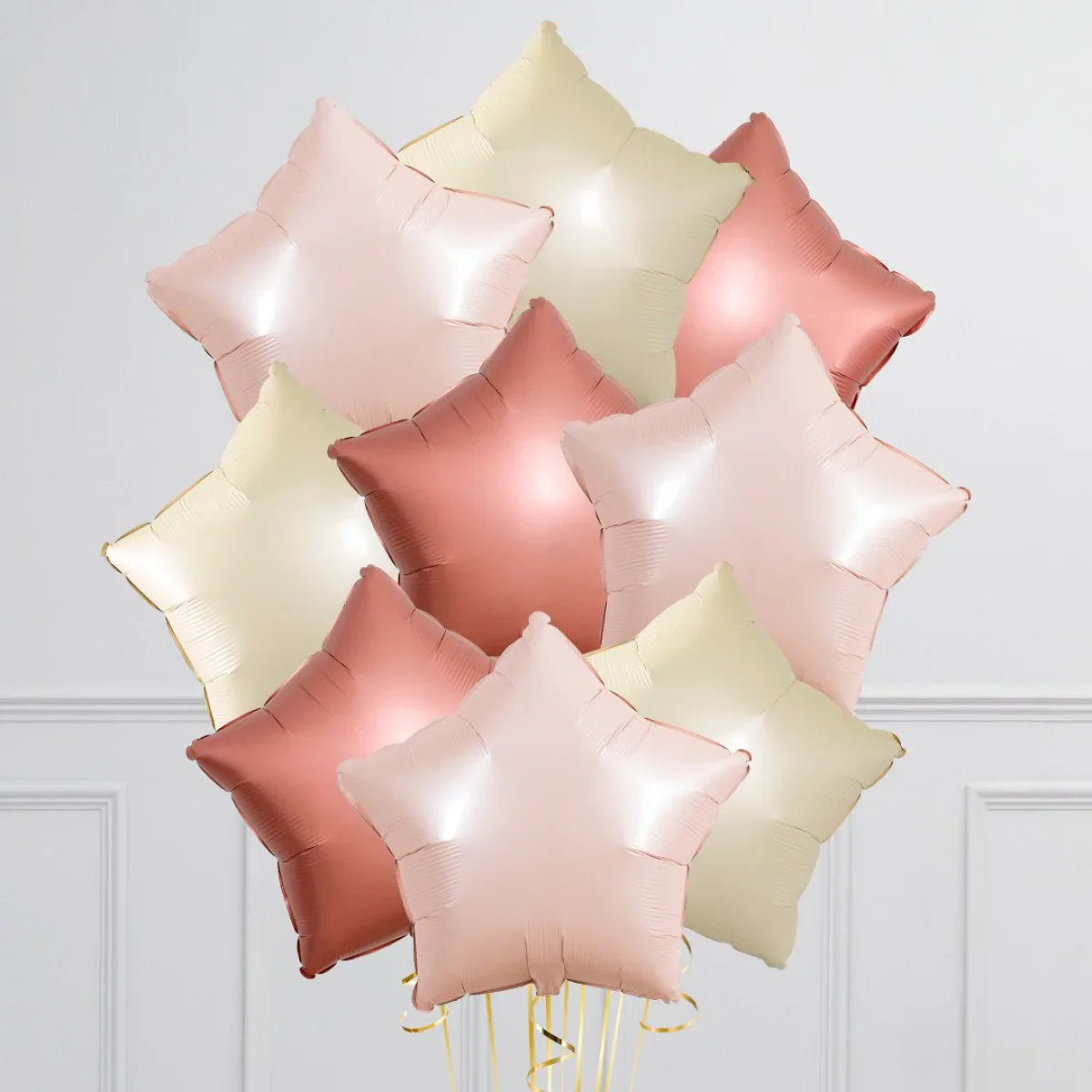 Cluster of metallic star-shaped foil balloons in soft pink, rose gold, and cream colors against white wall