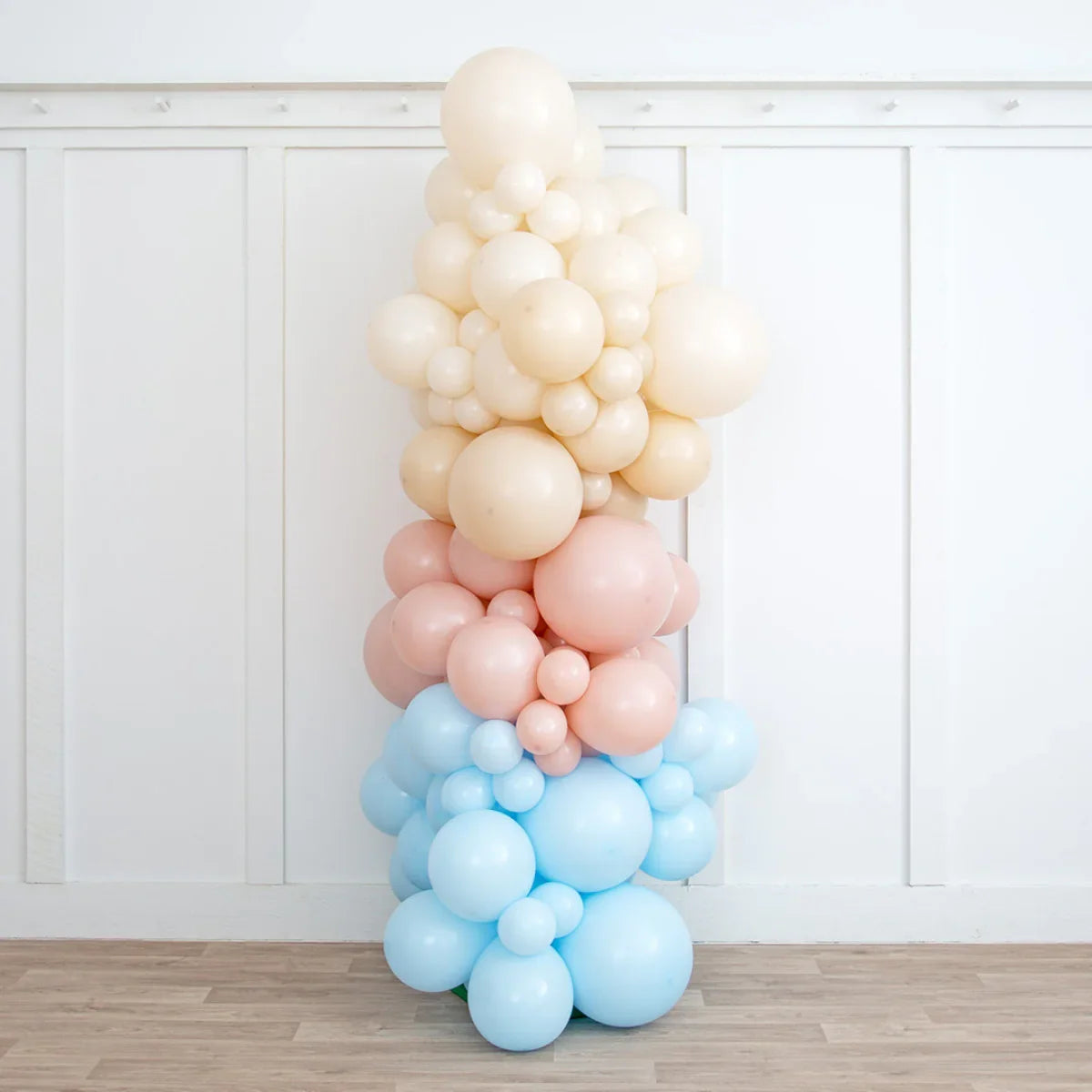 Neutral baby pink, cream, and blue balloon pillar decoration against white wall and wooden floor