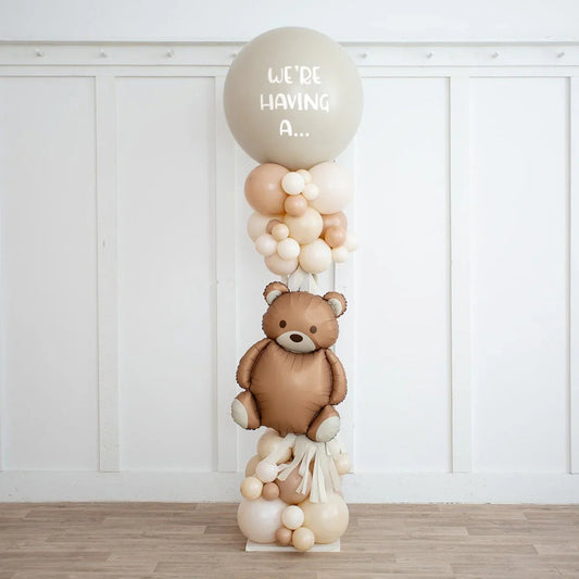 Neutral beige balloon pillar with teddy bear balloon and text we're having a for gender reveal party