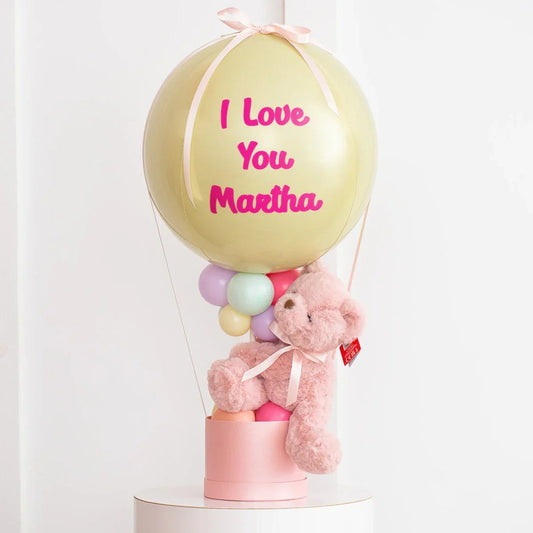 Pastel yellow balloon with pink text 'I Love You Martha' attached to pink box with pastel balloons and pink teddy bear
