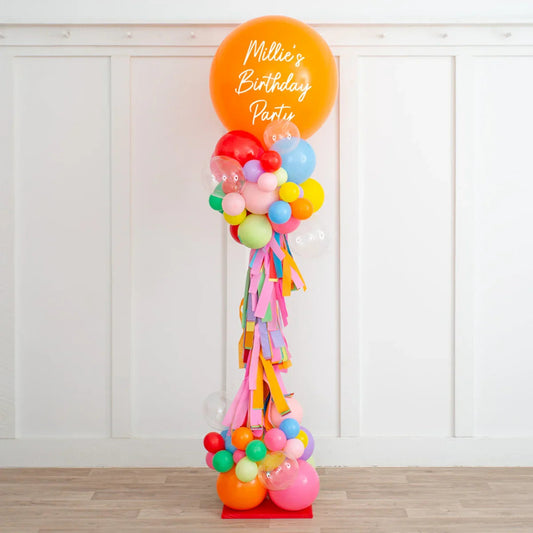 Tall colorful birthday balloon pillar with orange top balloon reading Millie's Birthday Party