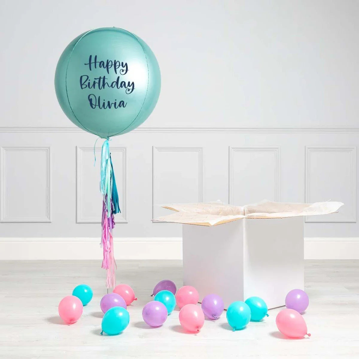 Teal inflated balloon with Happy Birthday Olivia text and colorful tassels next to an open white gift box and scattered small balloons