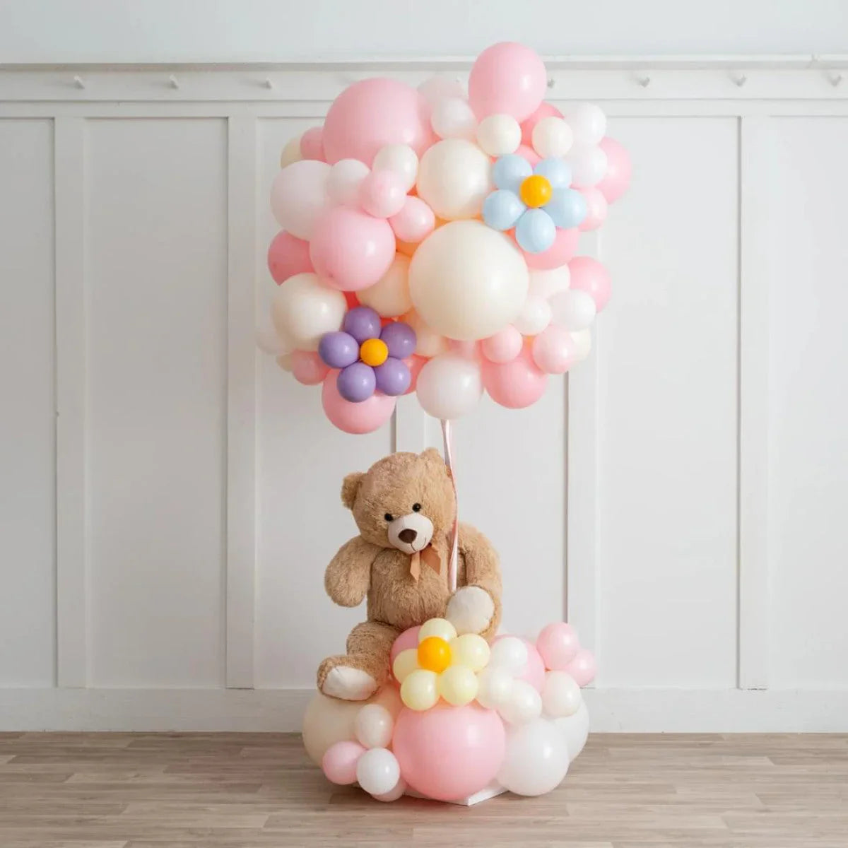 Teddy bear holding pastel pink and white balloon bouquet with flower-shaped balloons in a white room