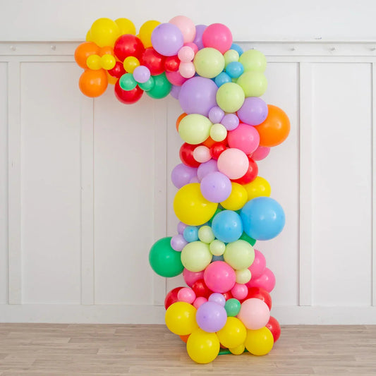 Asymmetric rainbow balloon arch decoration in pastel and bright colors against white paneled wall