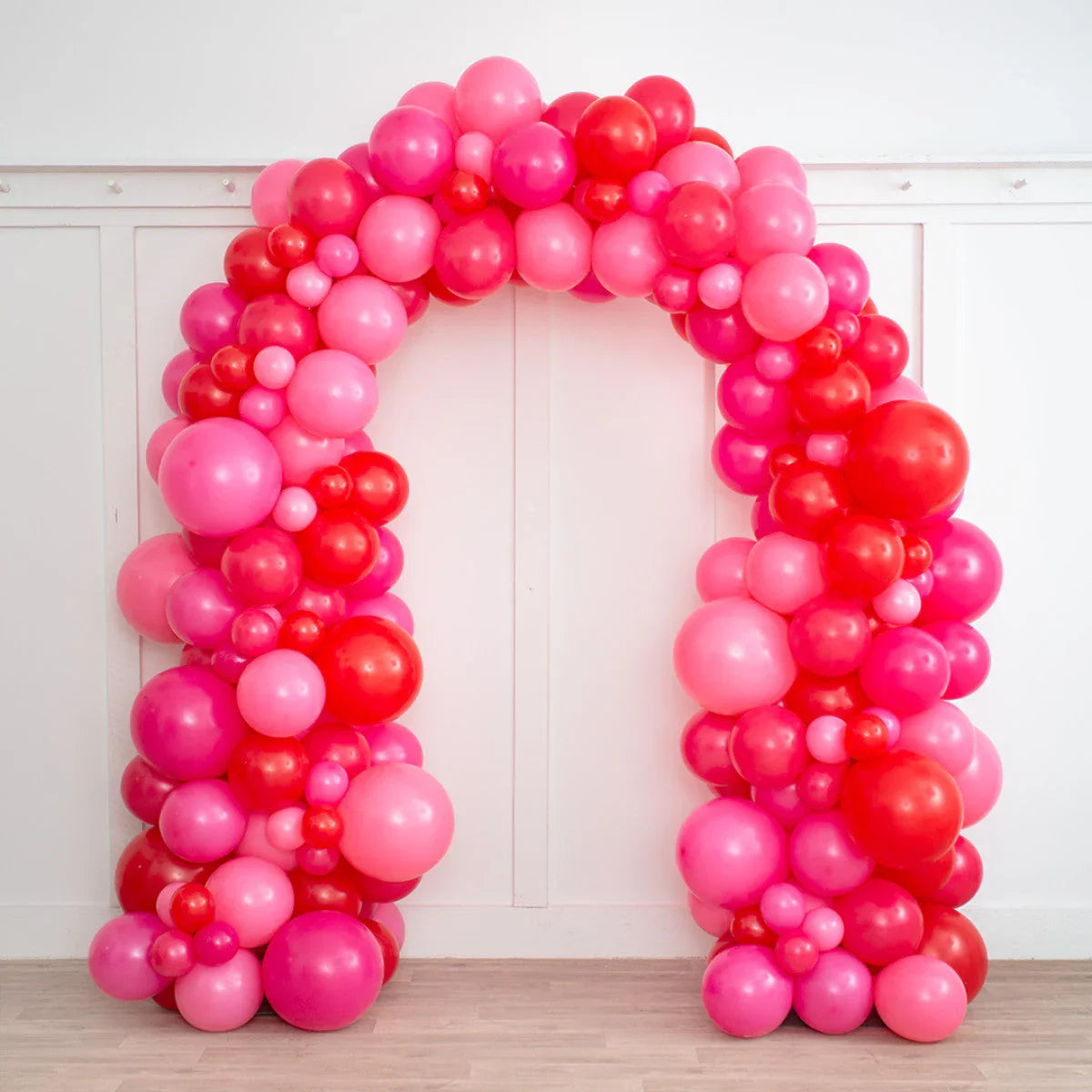 Raspberry red and pink balloon arch decoration with various balloon sizes, indoor setting with white wall and wooden floor