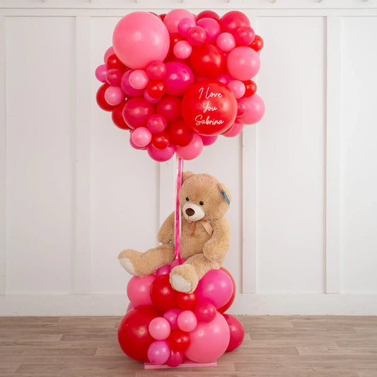 Teddy bear sitting among red and pink balloon clusters with one balloon reading I love you Sabrina