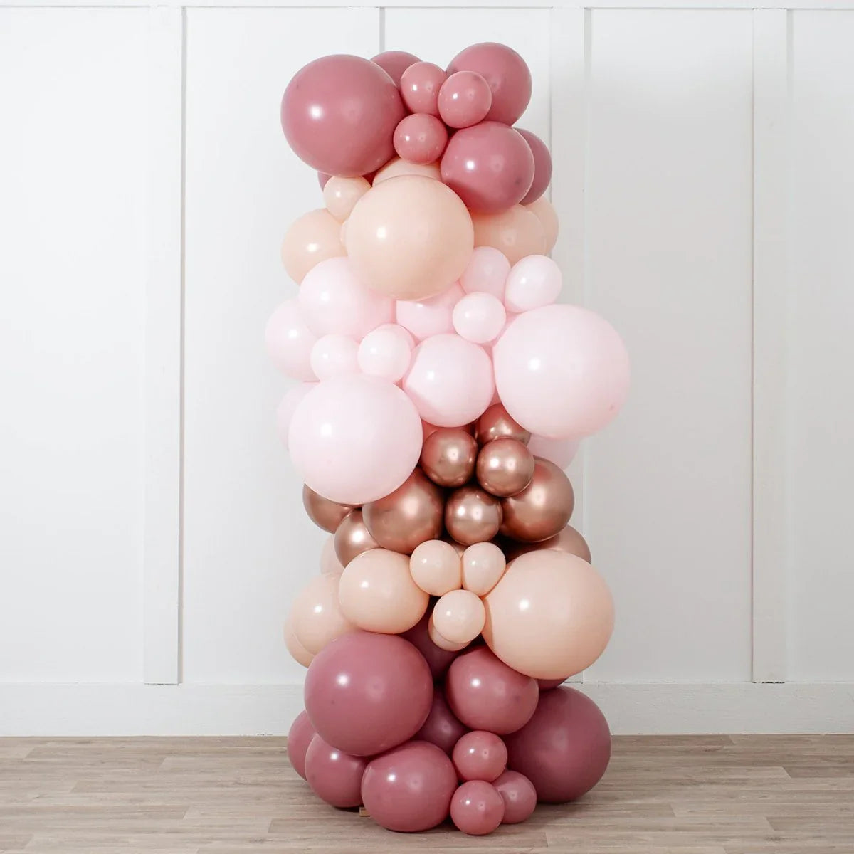 Rose gold, pink, and beige balloon pillar decoration against white wall on wooden floor