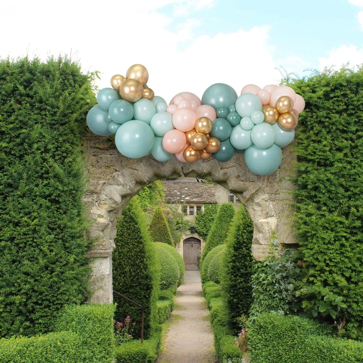 Sage green, blush pink, and gold balloon garland decorating stone archway in lush garden pathway