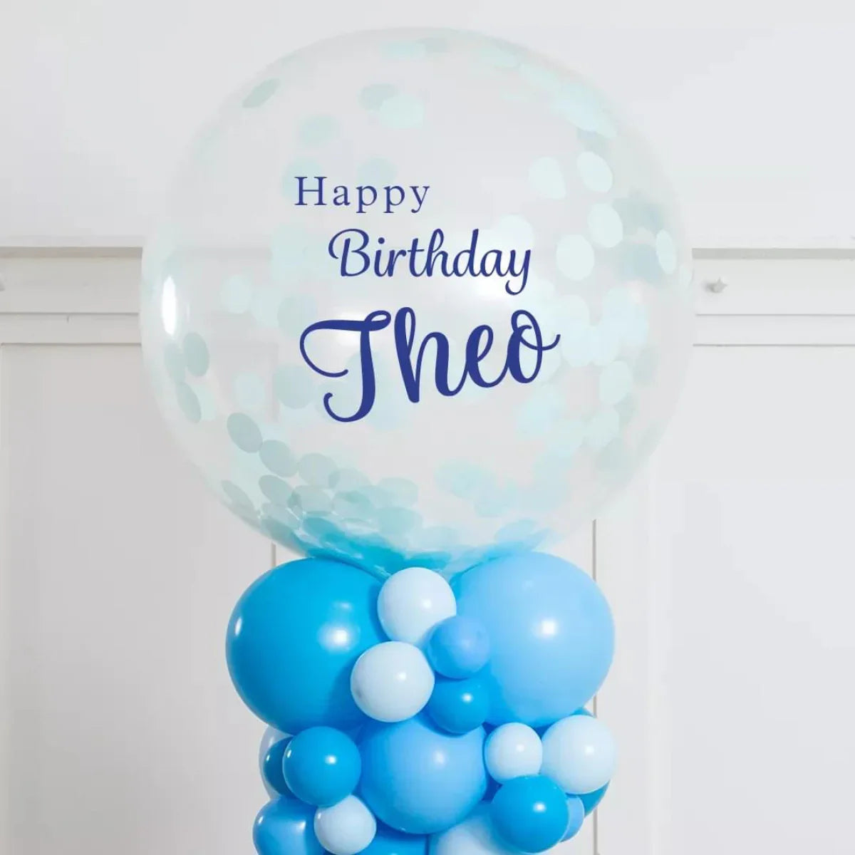 Transparent confetti balloon with Happy Birthday Theo text atop blue and light blue balloon pillar