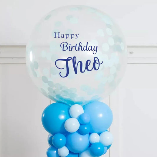 Transparent confetti balloon with Happy Birthday Theo text atop blue and light blue balloon pillar