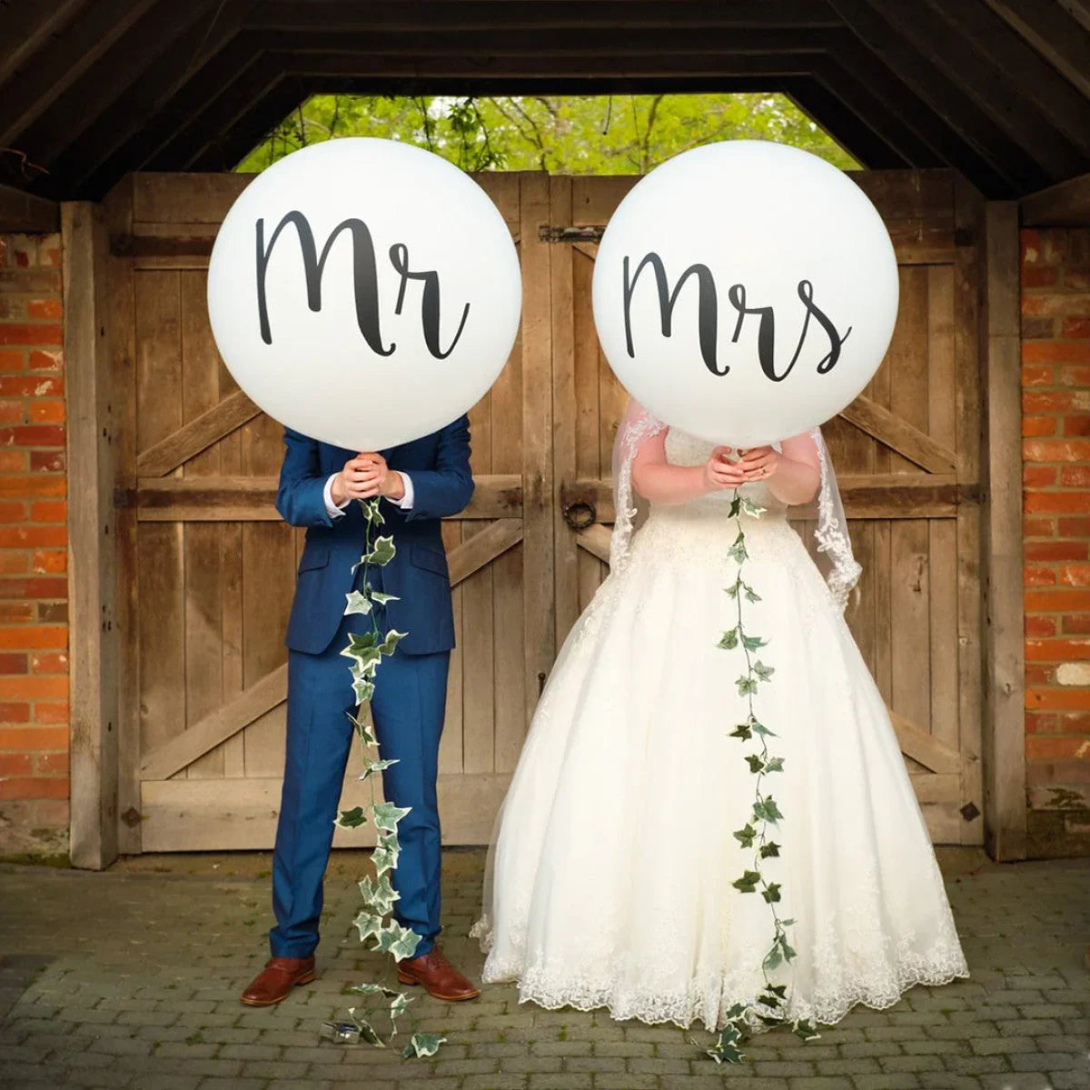 Bride and groom holding large white balloons with "Mr" and "Mrs" in front of rustic wooden barn doors