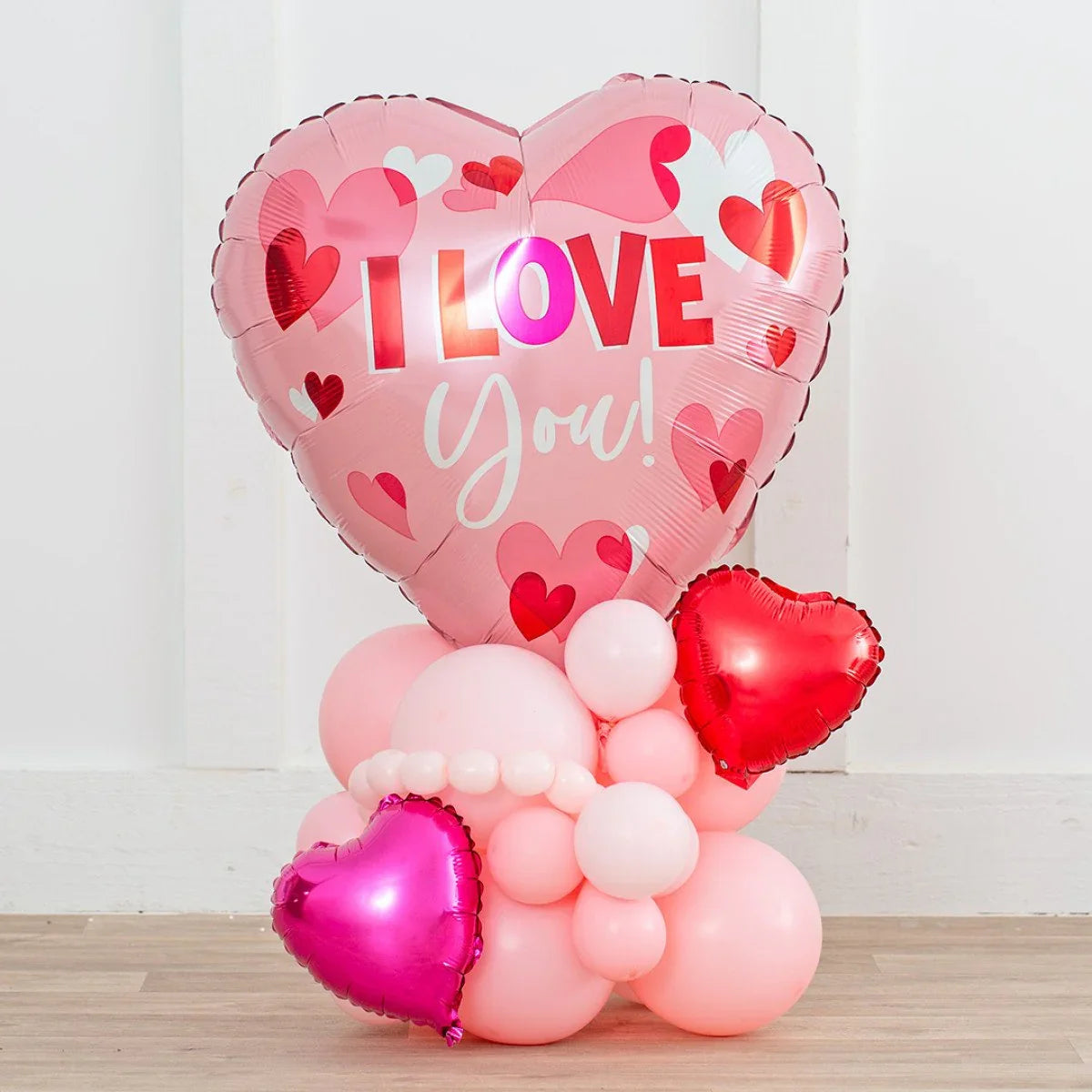 Pink heart-shaped balloon with I LOVE You text and smaller pink and red heart balloons on wooden floor