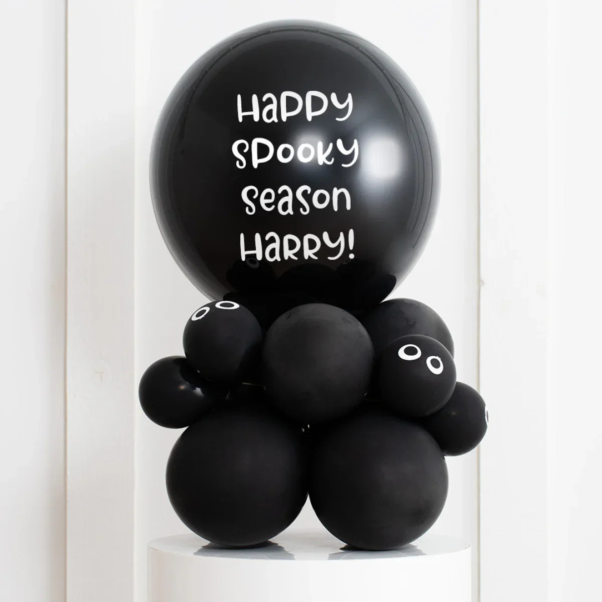 Black Halloween balloon arrangement with 'Happy spooky season Harry!' text and googly eyes on white pedestal