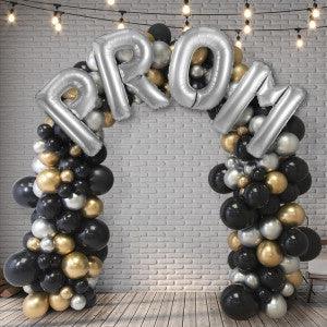 Silver PR0M balloon arch with black, gold, and silver balloons against white brick wall with string lights