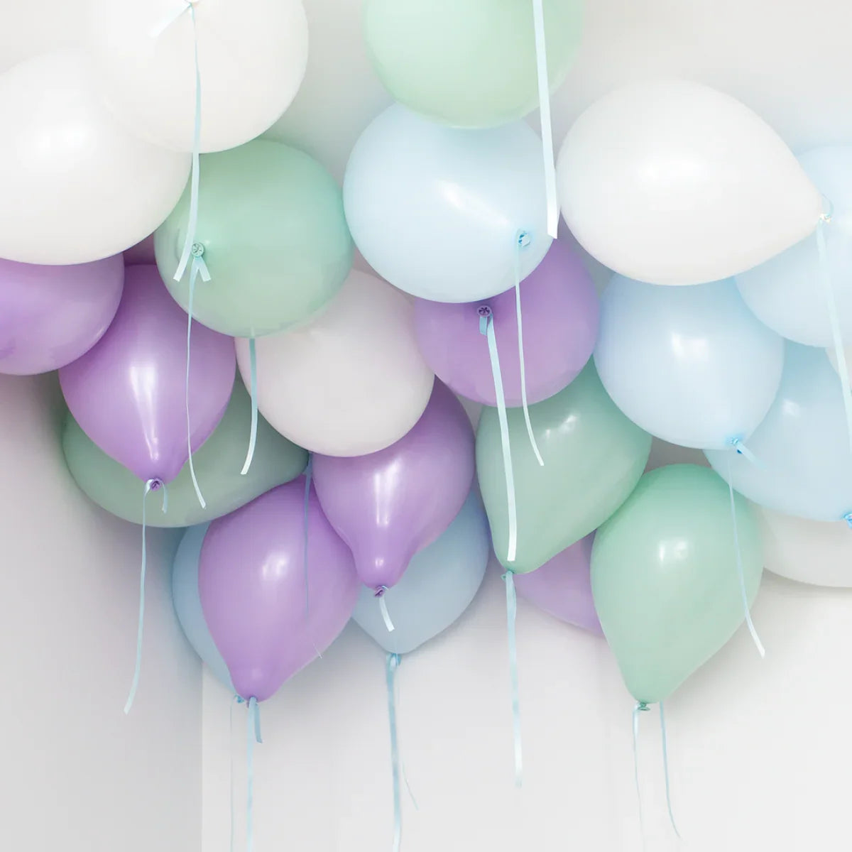 Cluster of pastel-colored helium balloons in purple, green, white, and light blue floating near a white ceiling corner
