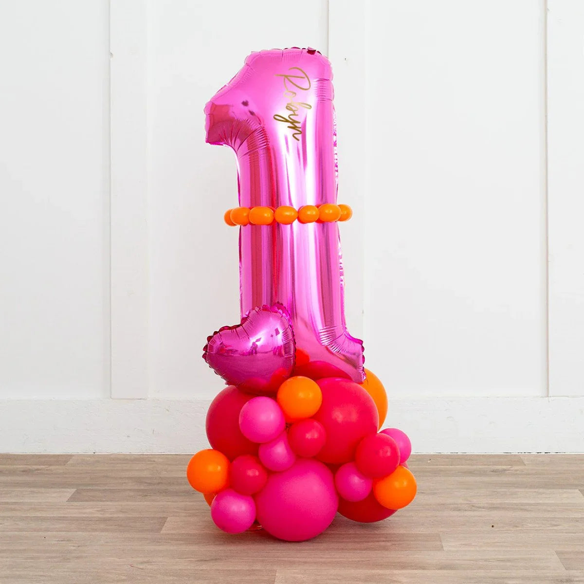 Pink foil balloon shaped as number 1 with orange and pink round balloons decoration on wood floor against white wall
