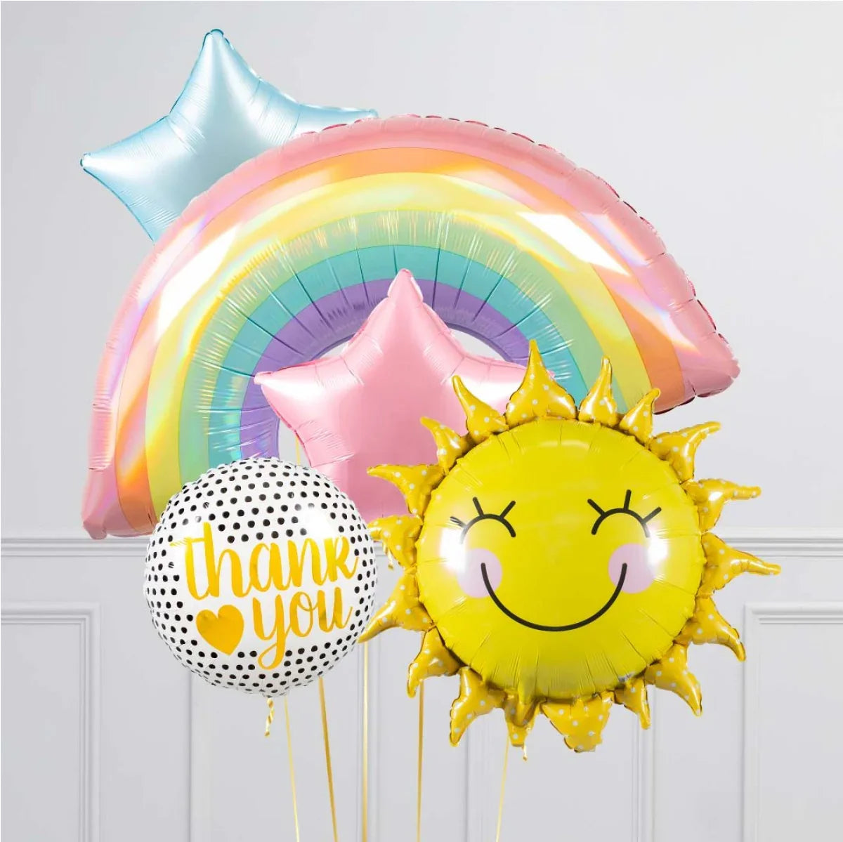 Colorful helium balloons including rainbow, smiling sun, pink star, and thank you balloons against white wall