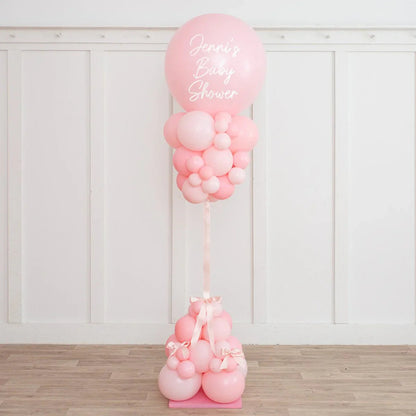 Pink baby shower balloon pillar with 'Jenni's Baby Shower' text in a white room