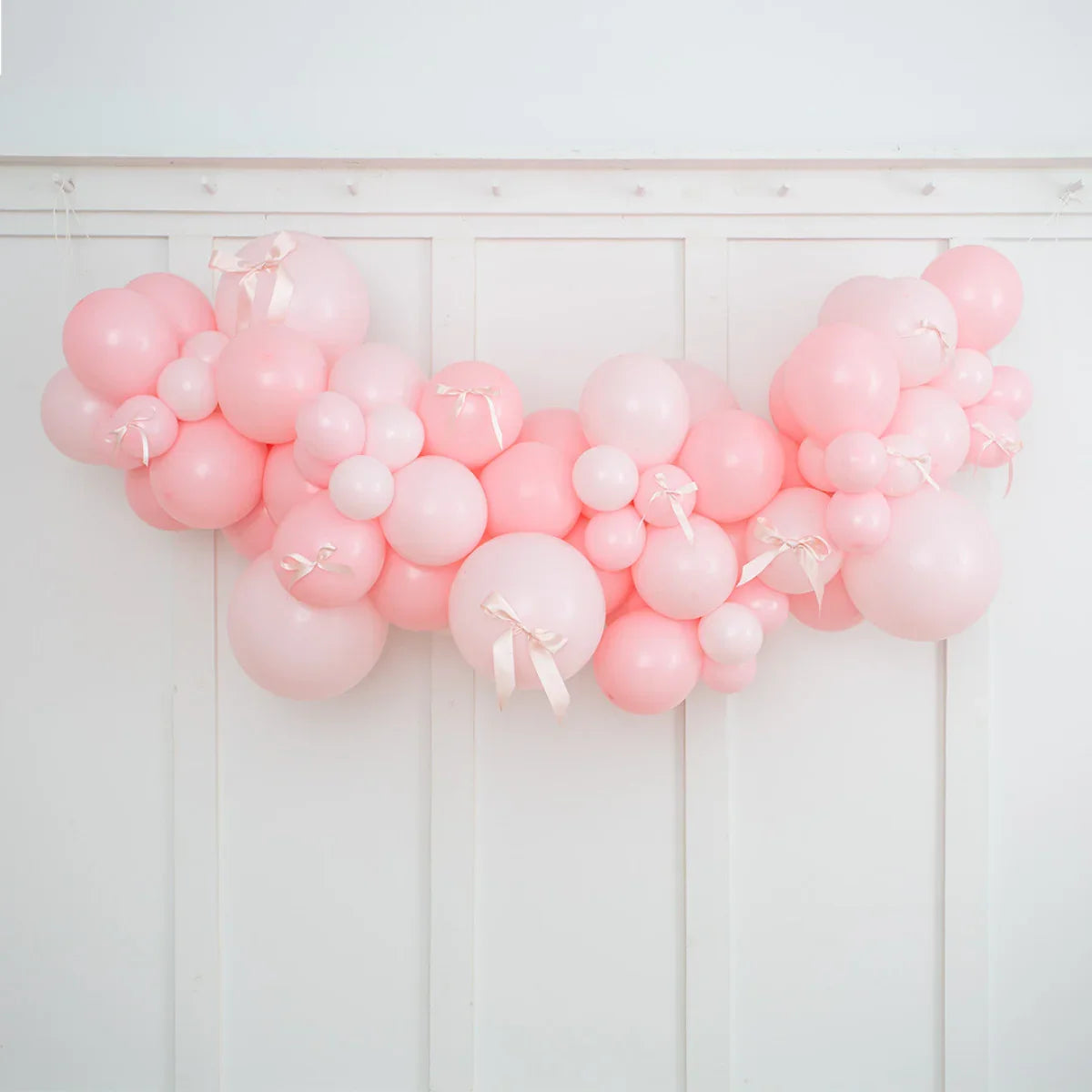 Baby pink and light pink balloon garland with satin bows hanging on white paneled wall