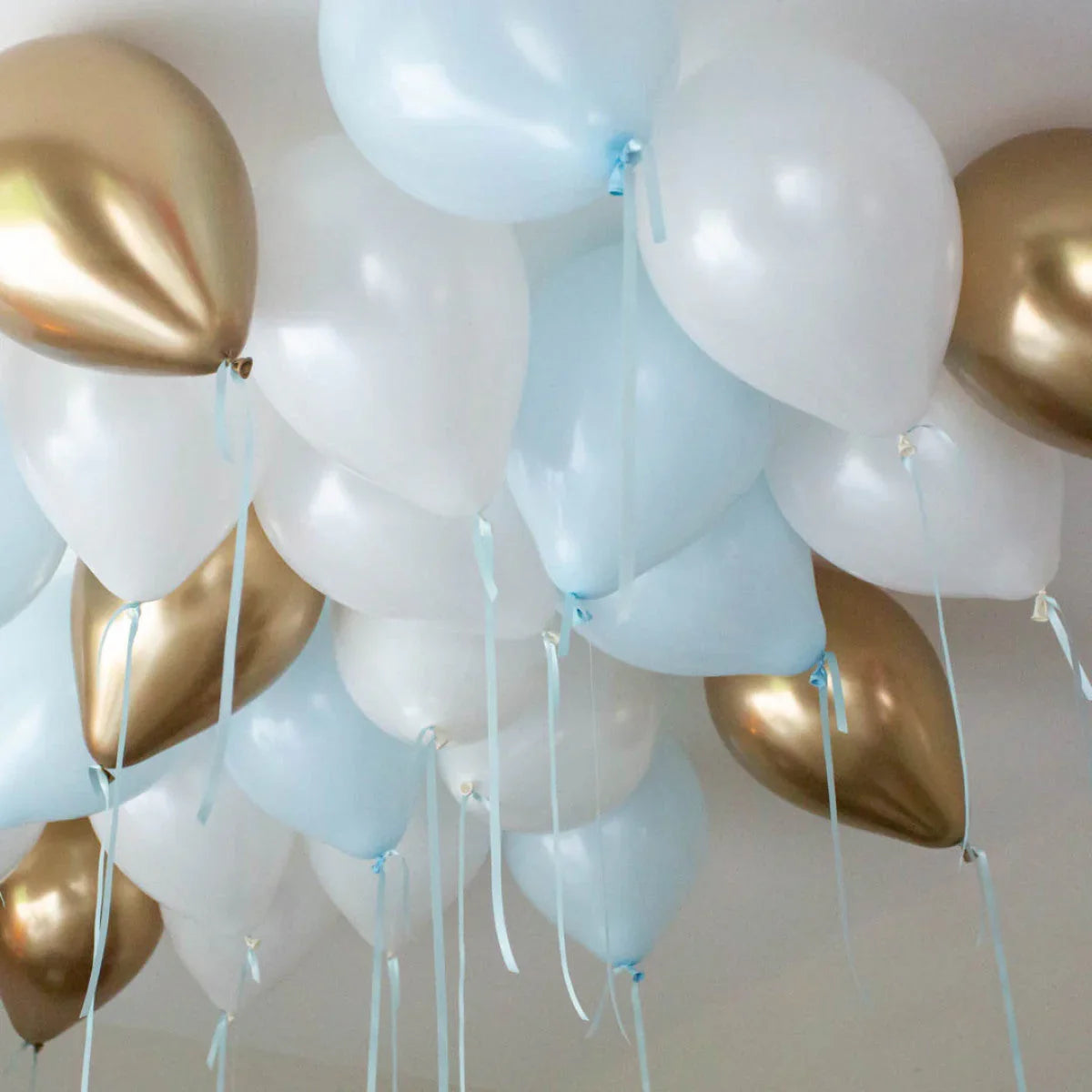 Cluster of white, light blue, and gold helium balloons floating on a ceiling with ribbons