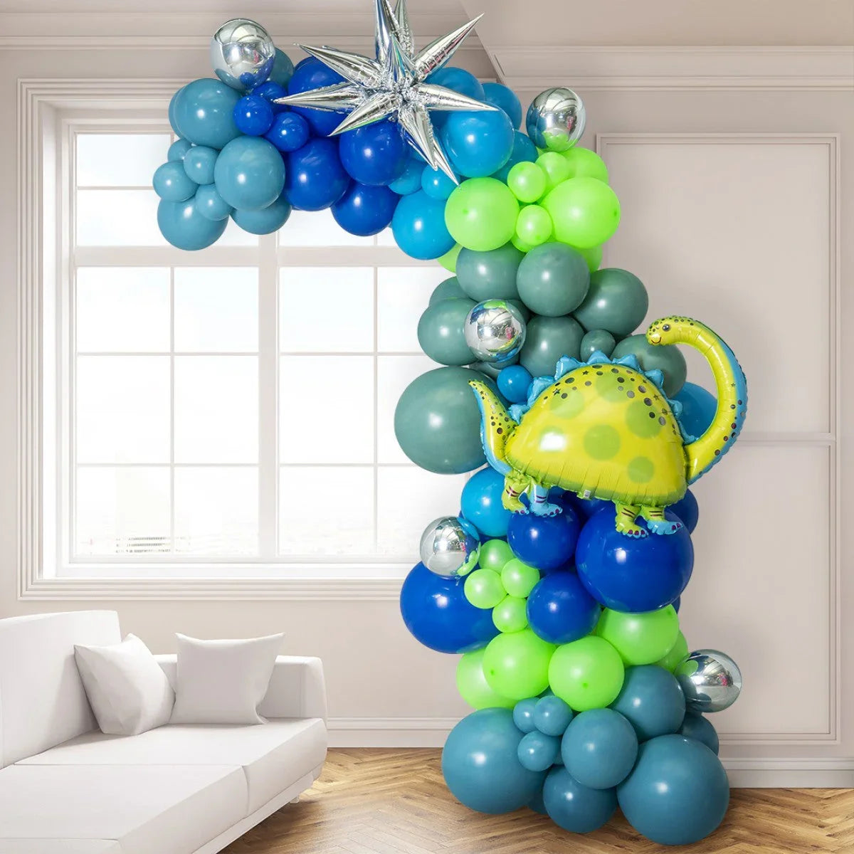 Asymmetric balloon arch with blue, green, and silver balloons featuring a yellow dinosaur foil balloon in a modern living room