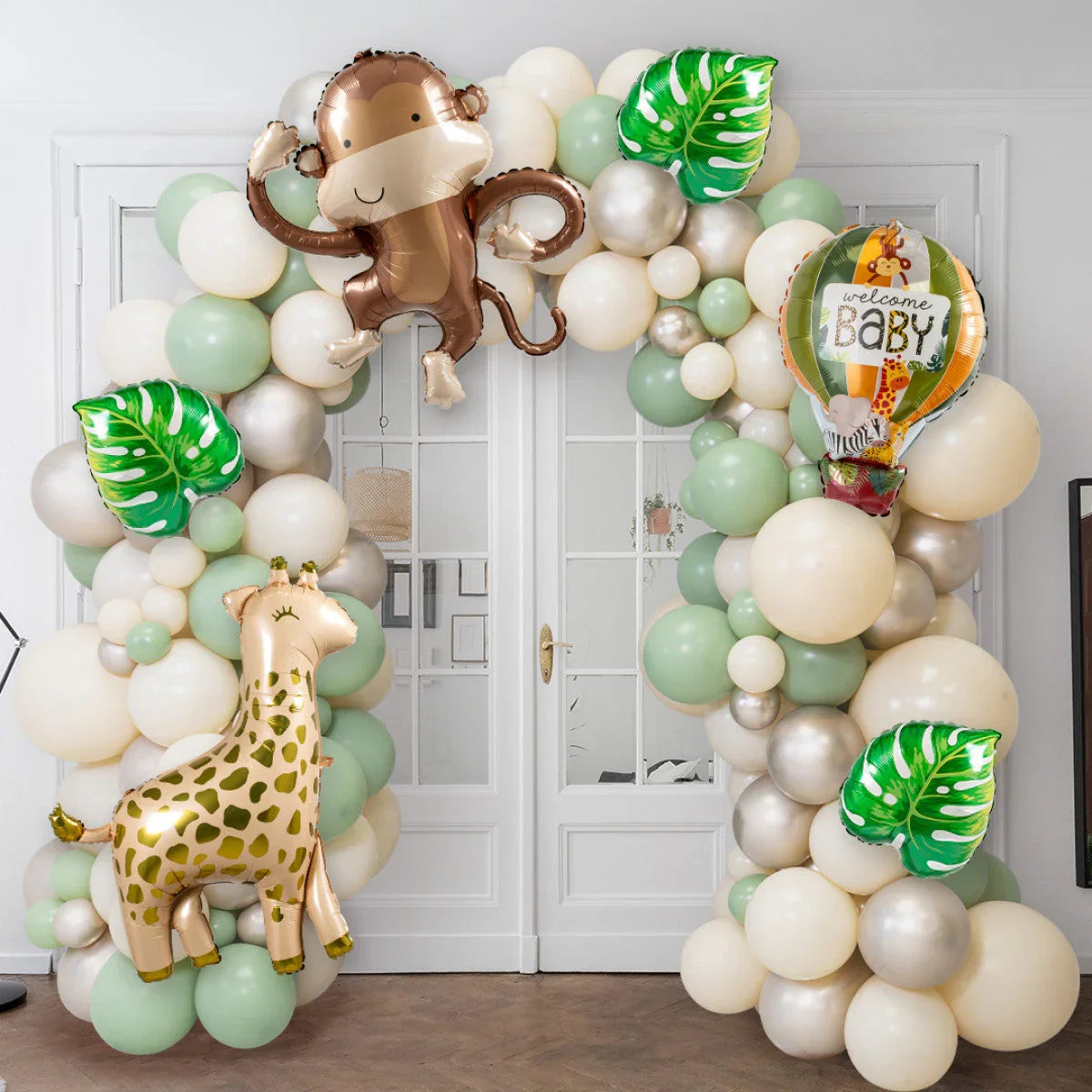 Jungle-themed baby shower balloon arch with green, cream, and gold balloons, monkey, giraffe, and welcome baby hot air balloon