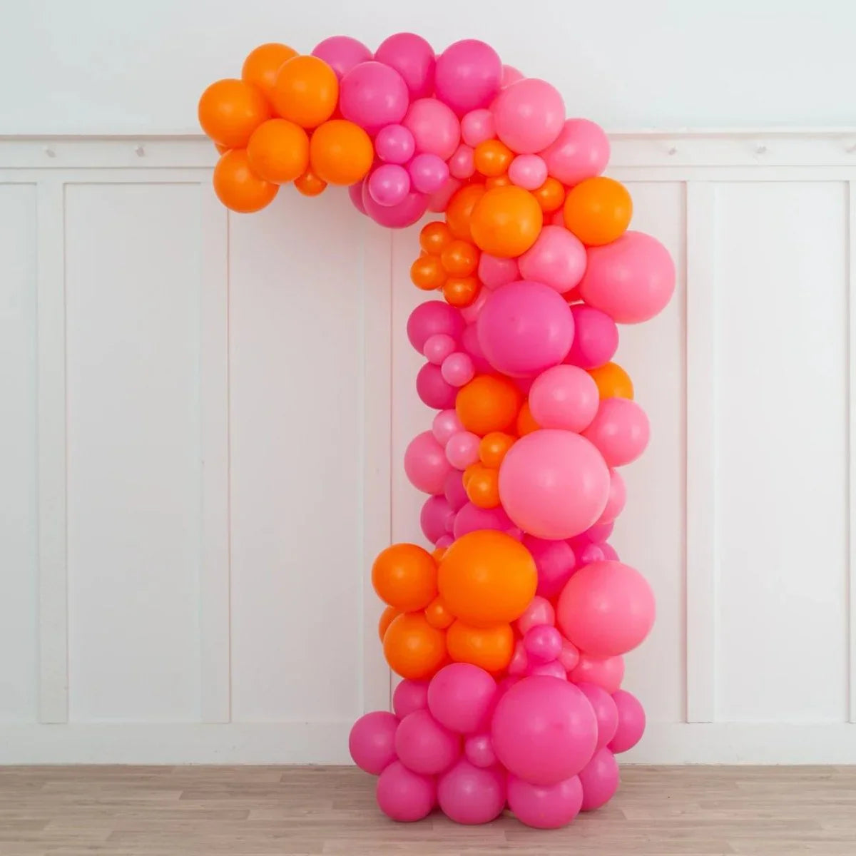 Asymmetric balloon arch in various shades of pink and orange against white paneled wall
