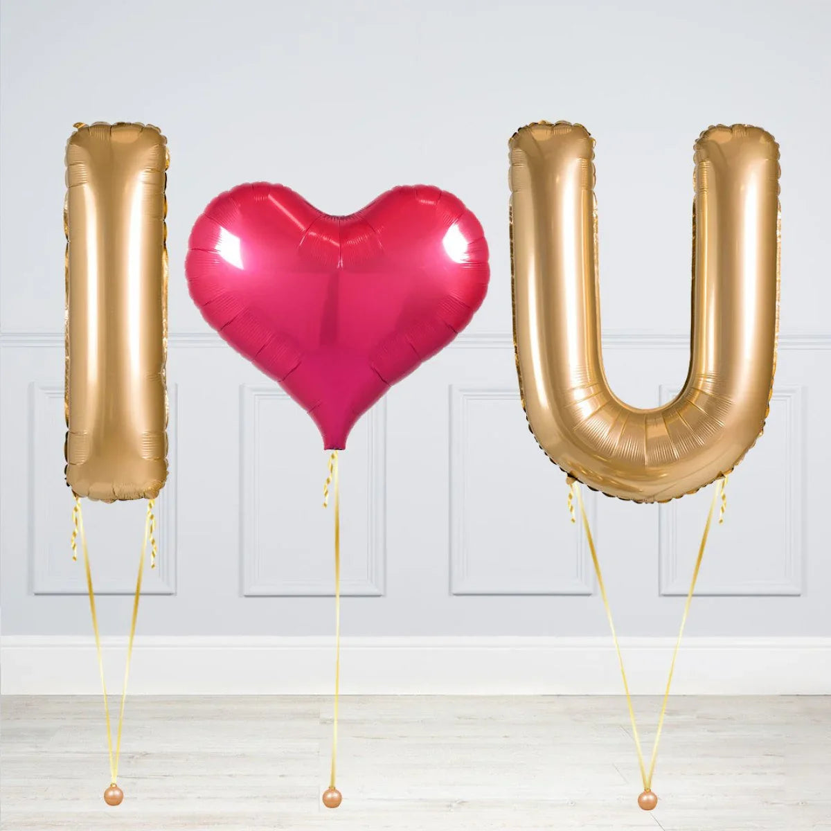 Gold foil balloons spelling I and U with a red heart-shaped balloon in between on a white wall background