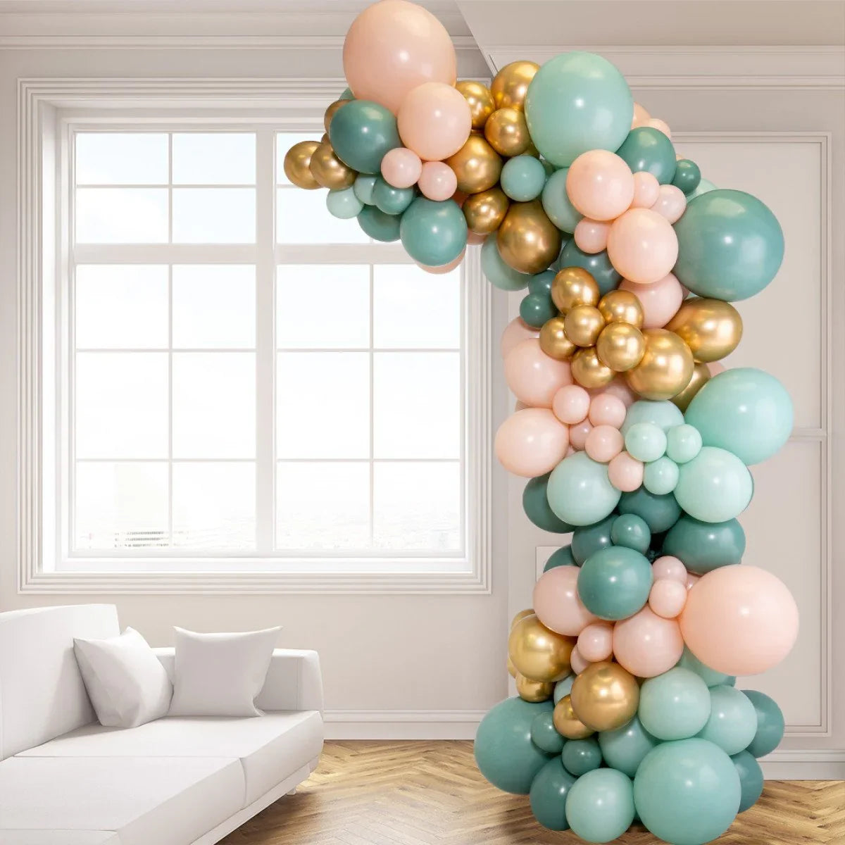 Asymmetric balloon arch in sage green, gold, and peach colors in a bright modern room with large window and white sofa