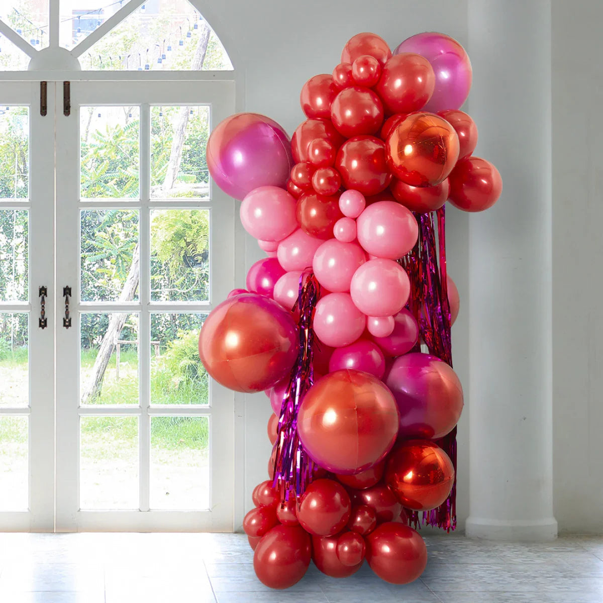 Red and pink chrome balloon pillar decoration indoors near white window with greenery outside