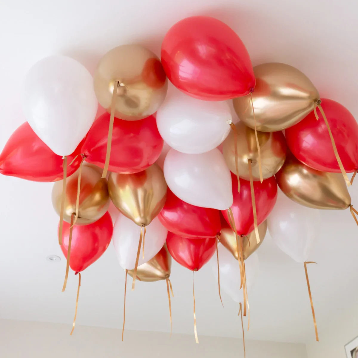 Cluster of red, white, and gold helium balloons floating near a white ceiling with gold ribbon tails