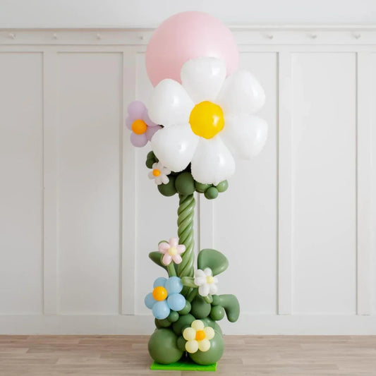 Tall balloon sculpture of a large white daisy with yellow center and smaller colorful flowers on wooden floor
