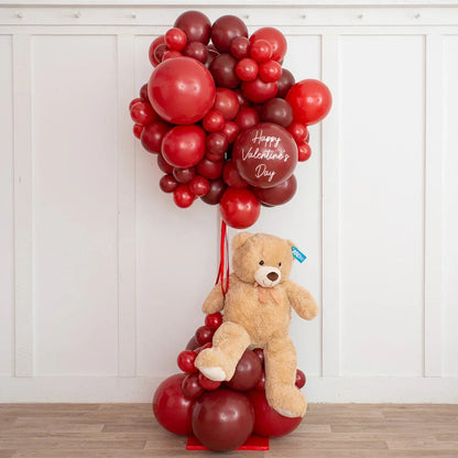 Red and dark cherry balloon bouquet with 'Happy Valentine's Day' balloon and beige teddy bear on wooden floor