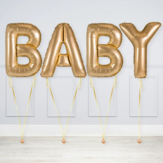 Gold foil letter balloons spelling BABY against a white paneled wall on wooden floor