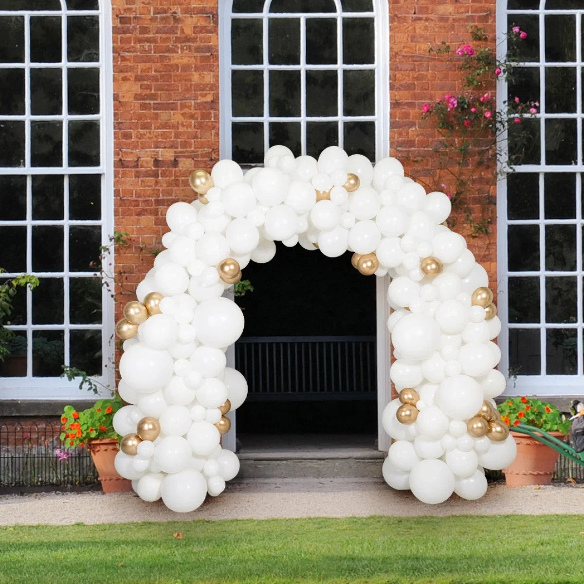 White and gold balloon arch decoration outside a brick building with large windows and potted flowers