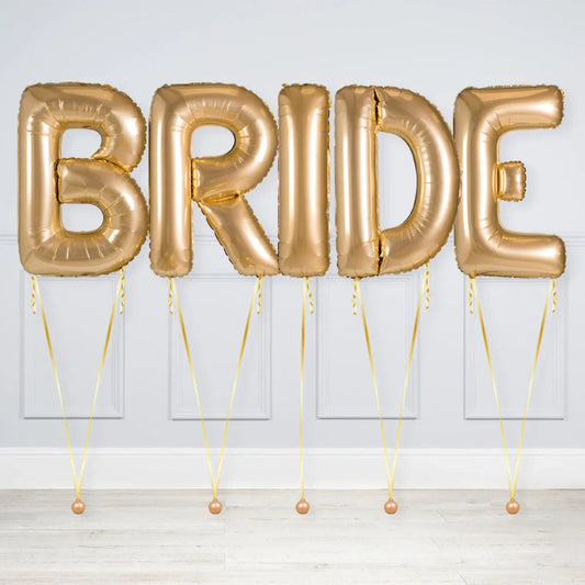 Gold foil balloon letters spelling BRIDE tied with ribbons on floor against gray wall