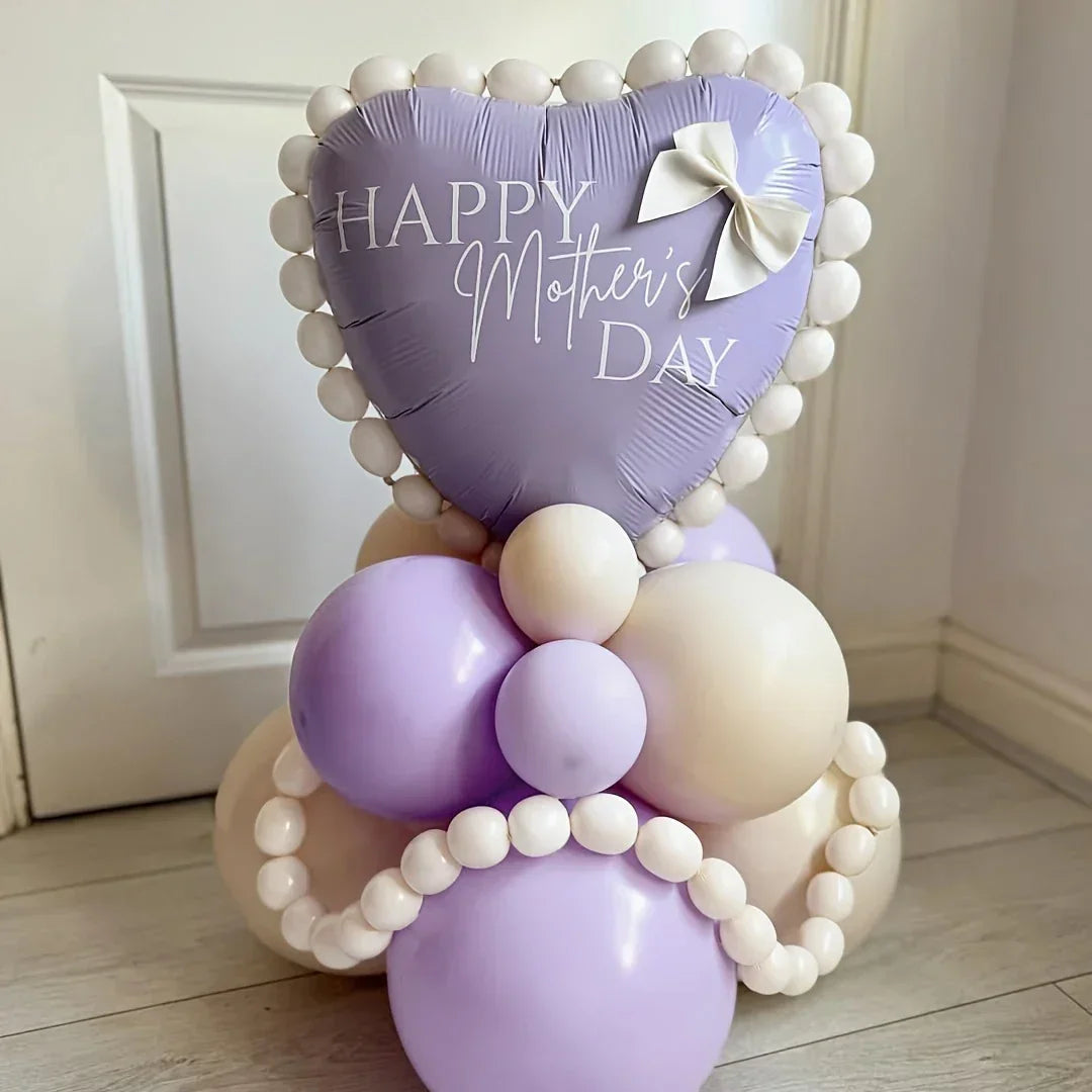 Heart-shaped purple balloon with 'Happy Mother's Day' text, surrounded by cream and purple balloons, indoor setting