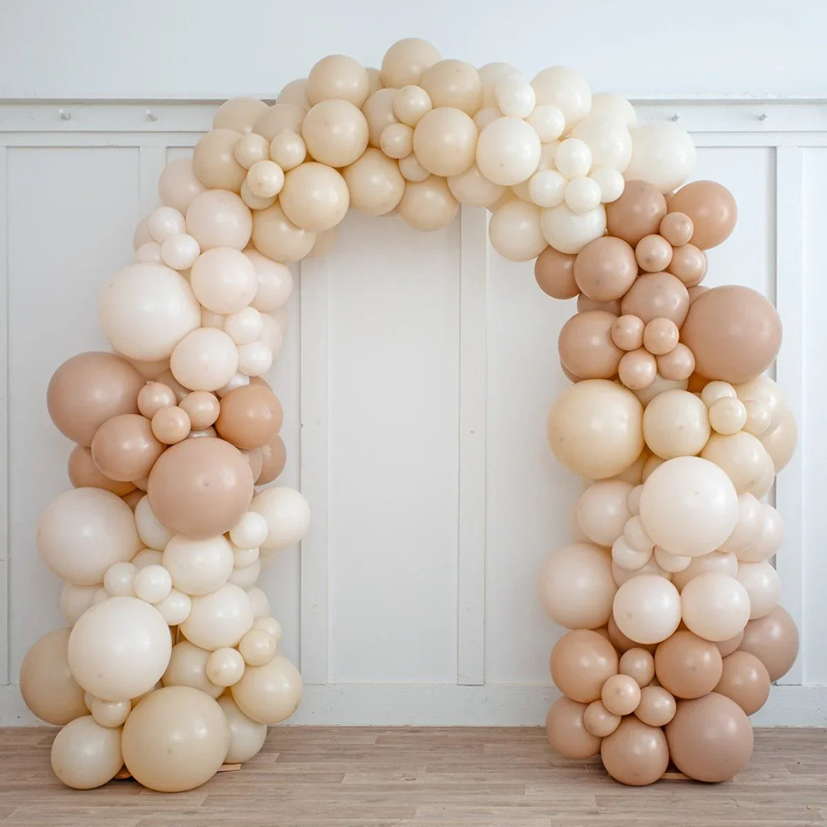 Neutral beige balloon arch with varying sizes set against white paneled wall on wooden floor