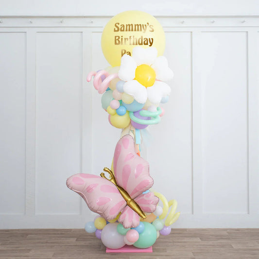 Personalised pastel balloon party pillar with pink butterfly, white daisy, and colorful balloons on wooden floor