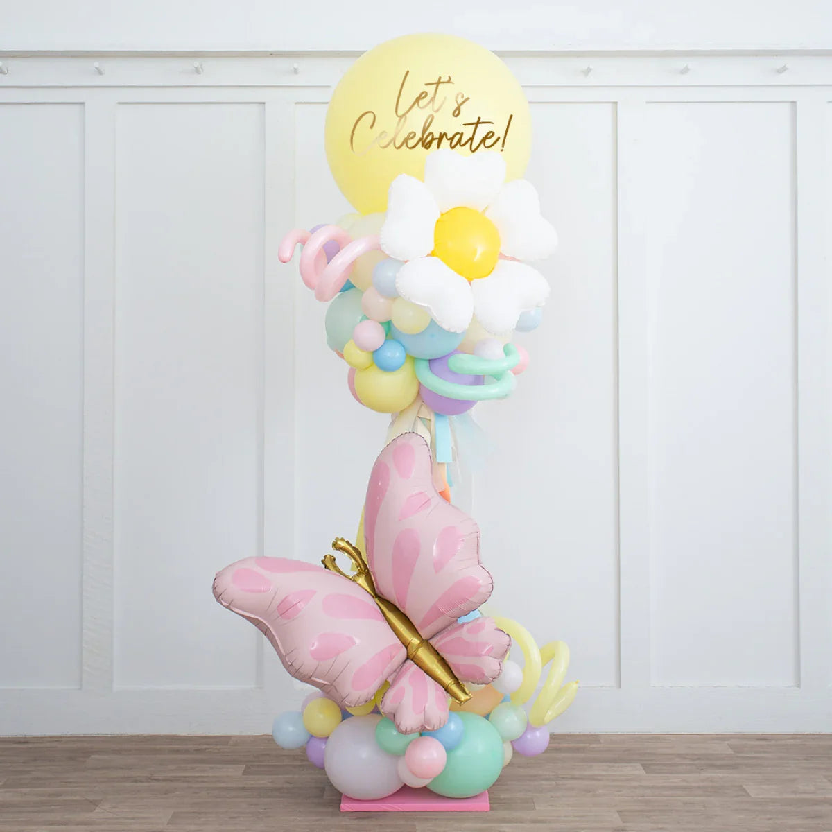 Pastel rainbow balloon pillar with pink butterfly, daisy flower, and yellow 'Let's Celebrate' balloon