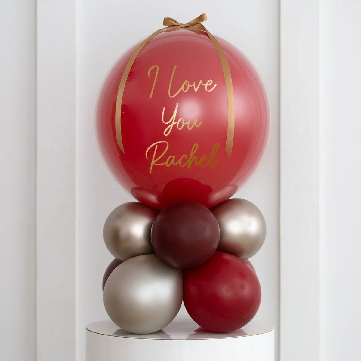 Personalized deep red pearl balloon stack with gold ribbon, reading 'I Love You Rachel', on white pedestal