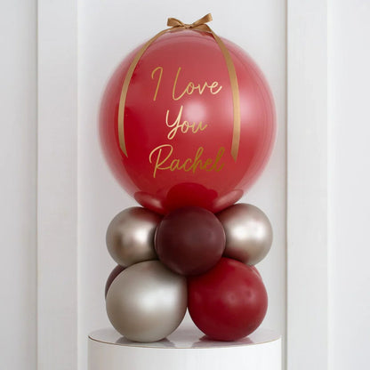 Personalized deep red pearl balloon stack with gold ribbon, reading 'I Love You Rachel', on white pedestal