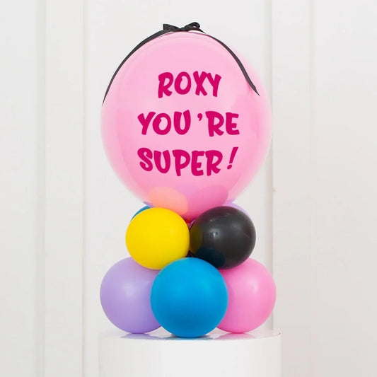 Personalised pink air-filled bubble balloon with message 'Roxy you're super!' on colorful balloon stack