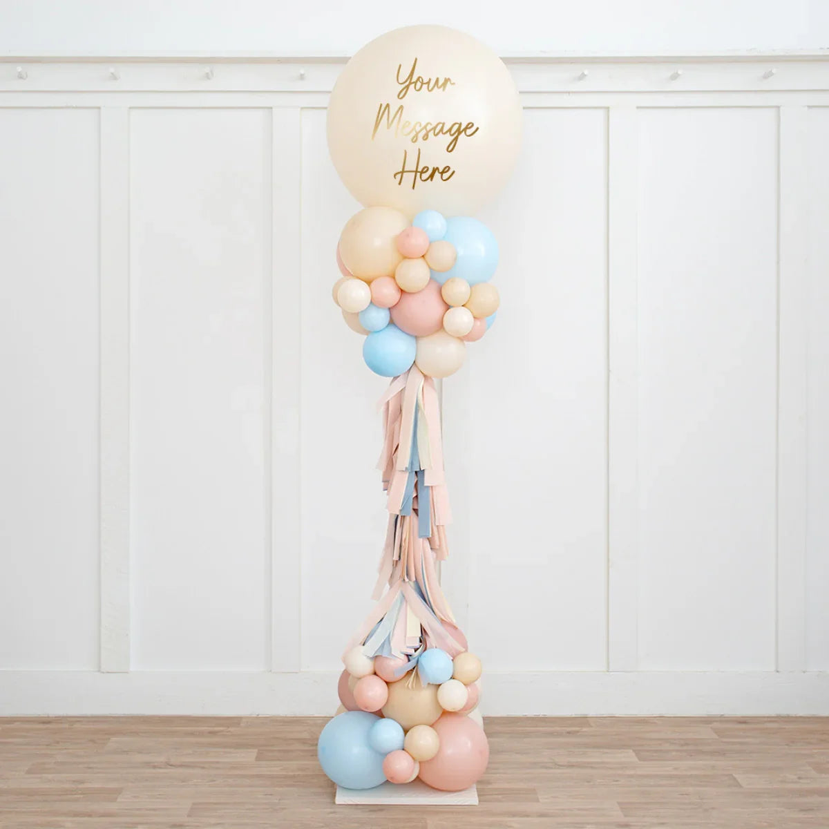 Tall balloon pillar with personalized message in beige, baby pink, and blue colors, indoor wooden floor